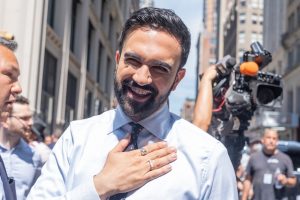 Zohran Mamdani souriant en public, main sur le plexus, entouré de caméras à New York, il est sorti victorieux de la primaire démocrate de 2025