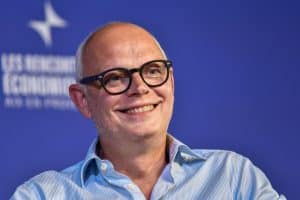 Homme souriant portant des lunettes noires et une chemise bleue à rayures, devant le fond bleu des Rencontres économiques