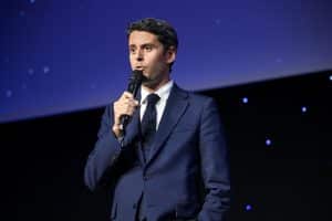 Homme en costume bleu marine et cravate à pois parlant dans un micro sur une scène éclairée de bleu