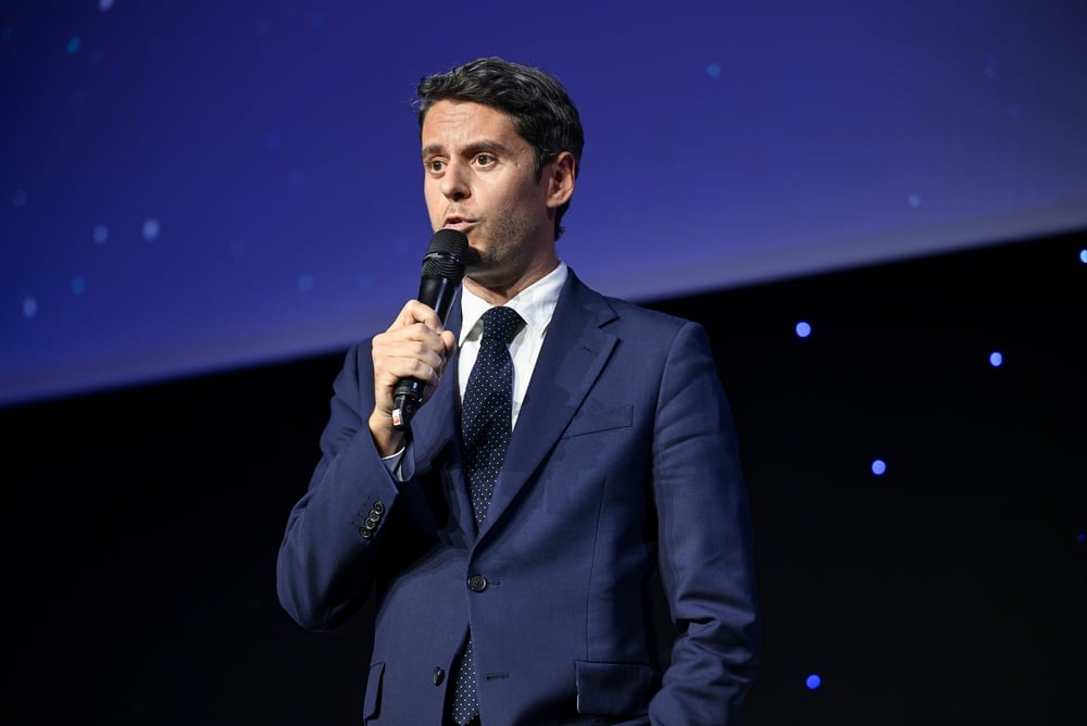 Homme en costume bleu marine et cravate à pois parlant dans un micro sur une scène éclairée de bleu