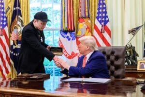 Poignée de main dans le Bureau ovale de la Maison-Blanche, entre un homme en casquette noire et un homme en costume bleu, devant des drapeaux américains